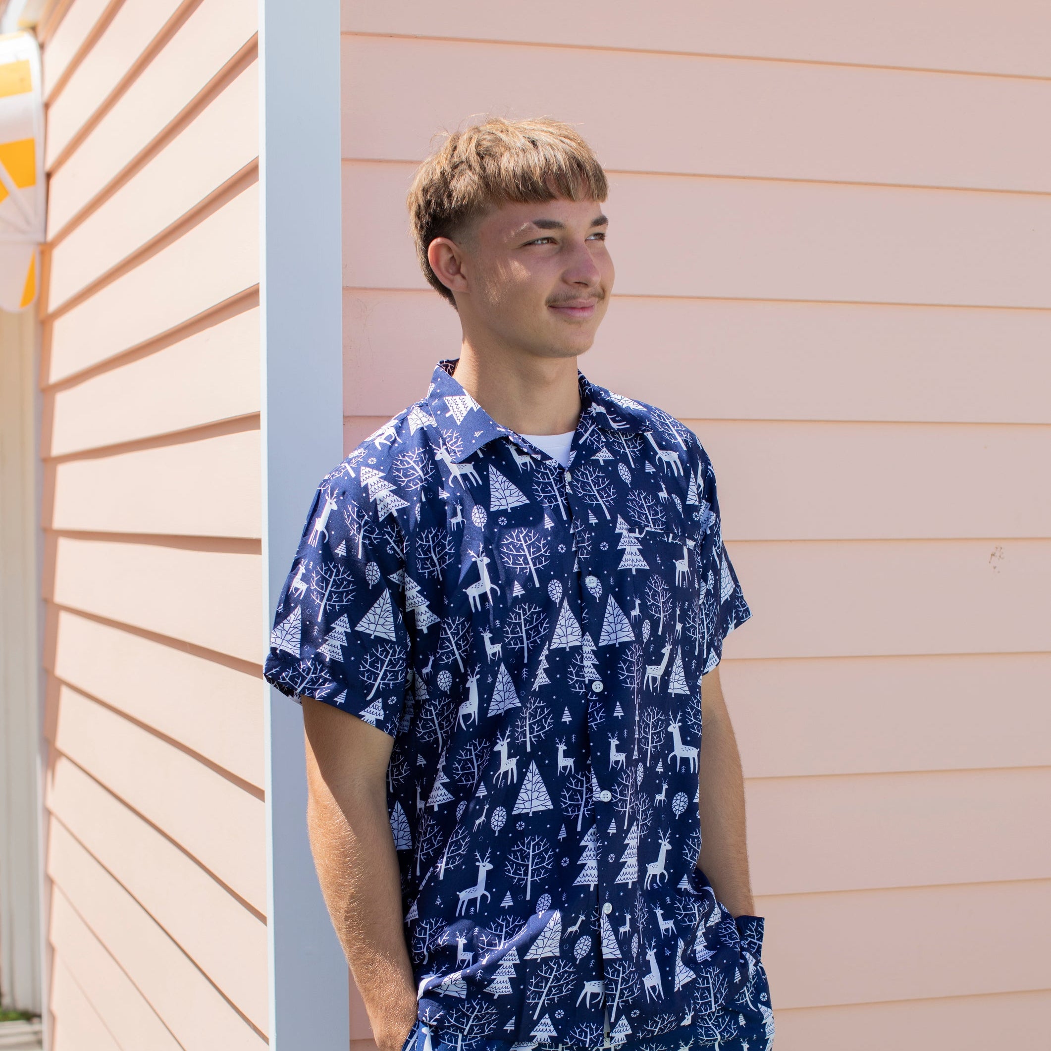 A young man wearing a blue and white patterned short-sleeve button-up shirt and matching shorts, standing outdoors against a peach-colored house siding.