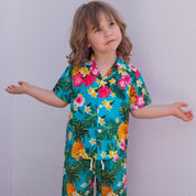 A young child wearing a vibrant tropical print short-sleeve pajama set, standing barefoot on a wooden deck against a white wall.