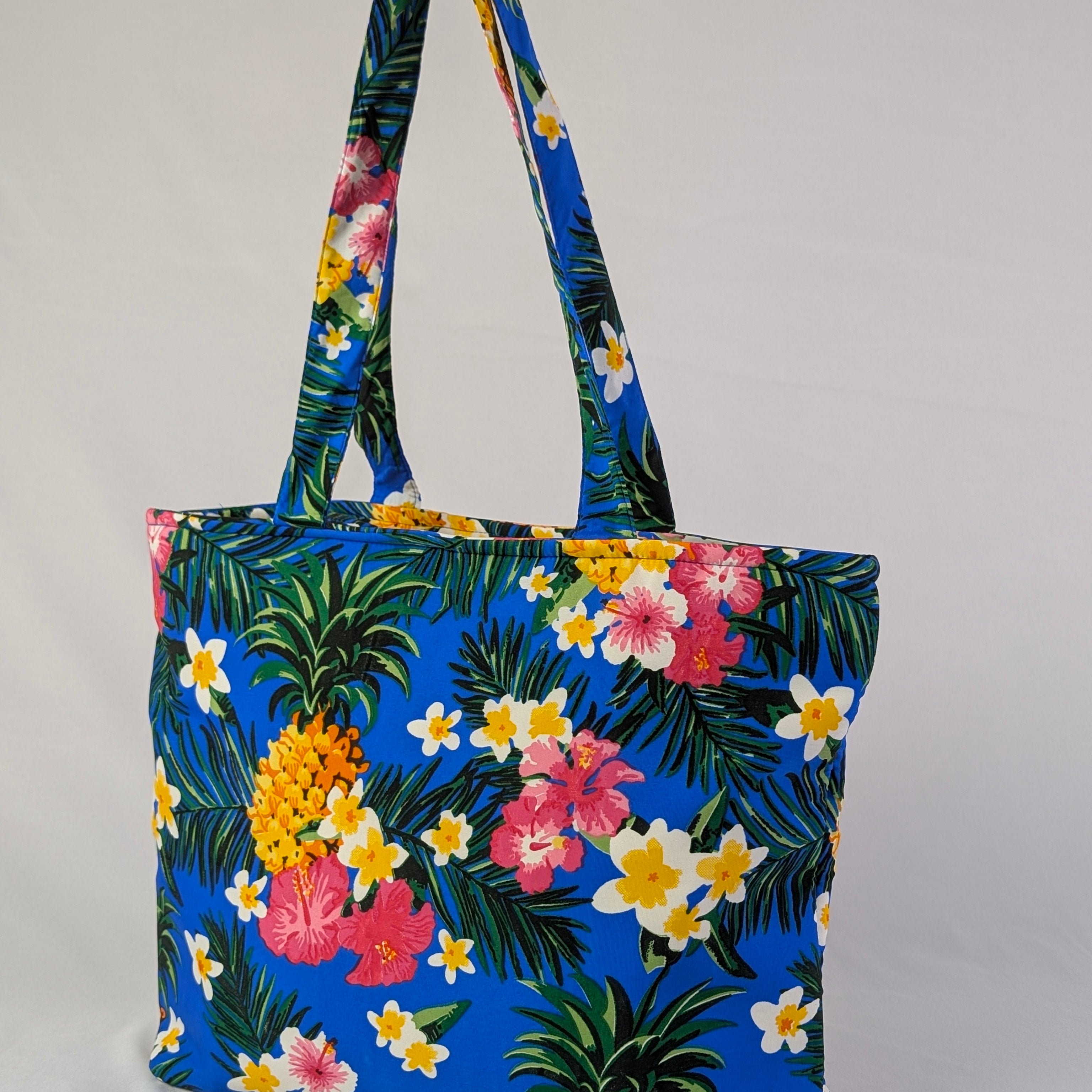 Blue tote bag with pineapple print and pink, yellow, and white tropical flowers; festive summer style. | Tropicool Clothing