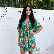 A woman wearing a vibrant green tropical floral print kaftan dress, standing barefoot on a stone patio in front of a white wooden fence.