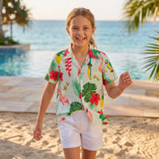 A young girl smiling on a sandy beach wearing a short-sleeve tropical print button-up shirt and white shorts, with a swimming pool and ocean in the background.
