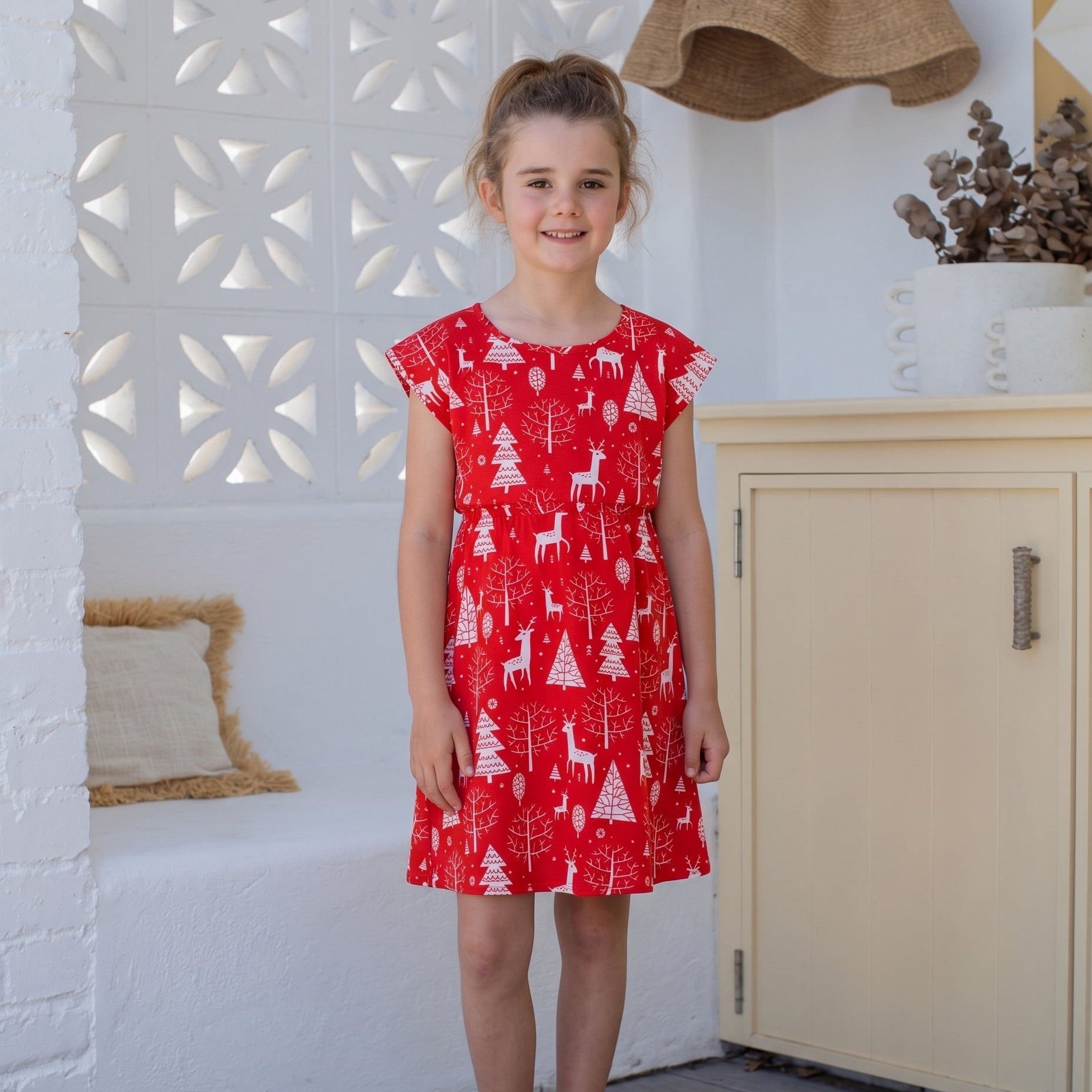 A young girl stands in a bright room wearing a red sleeveless dress with a white Christmas tree and deer pattern. She is wearing pink sandals and standing in front of a white decorative wall, a cream cabinet, and a wooden floor.