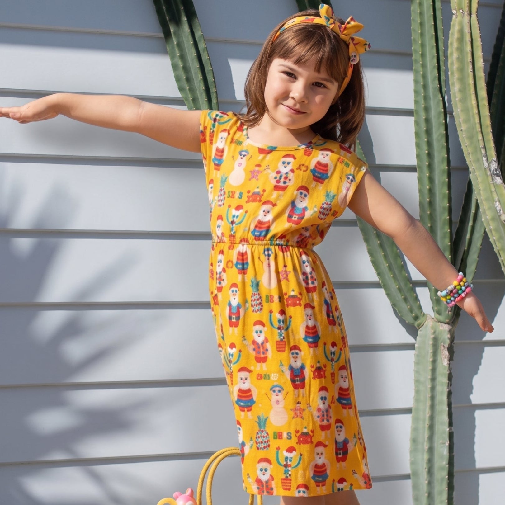 A young girl wearing a yellow short-sleeve dress with a colorful cartoon pattern, standing outdoors in front of a white slatted wall with cacti. She is holding a woven straw tote bag and wearing yellow sandals with floral details.