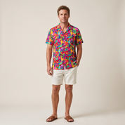 A man modeling a vibrant, multicolor tropical print short-sleeve button-down shirt paired with off-white shorts and brown sandals, standing against a solid light beige studio background.