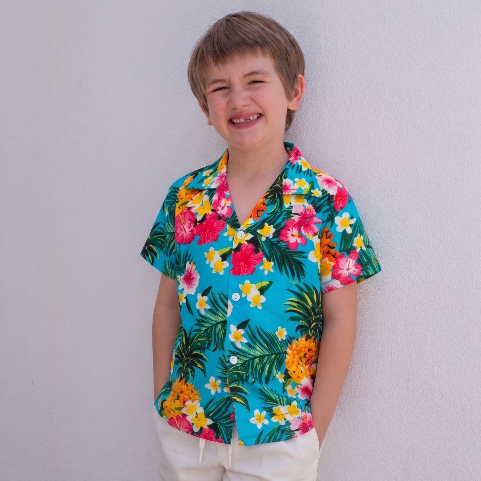 A young boy smiling and leaning against a white wall, wearing a vibrant turquoise short-sleeve button-up shirt with a colorful tropical floral and palm leaf print, paired with cream shorts.