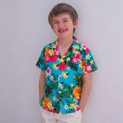A young boy smiling and leaning against a white wall, wearing a vibrant turquoise short-sleeve button-up shirt with a colorful tropical floral and palm leaf print, paired with cream shorts.