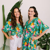 Two women modeling matching tropical print outfits in a bright indoor setting with potted plants. The women are wearing matching turquoise green short-sleeve button-up shirts and a matching V-neck dress with a pineapple and hibiscus flower print.