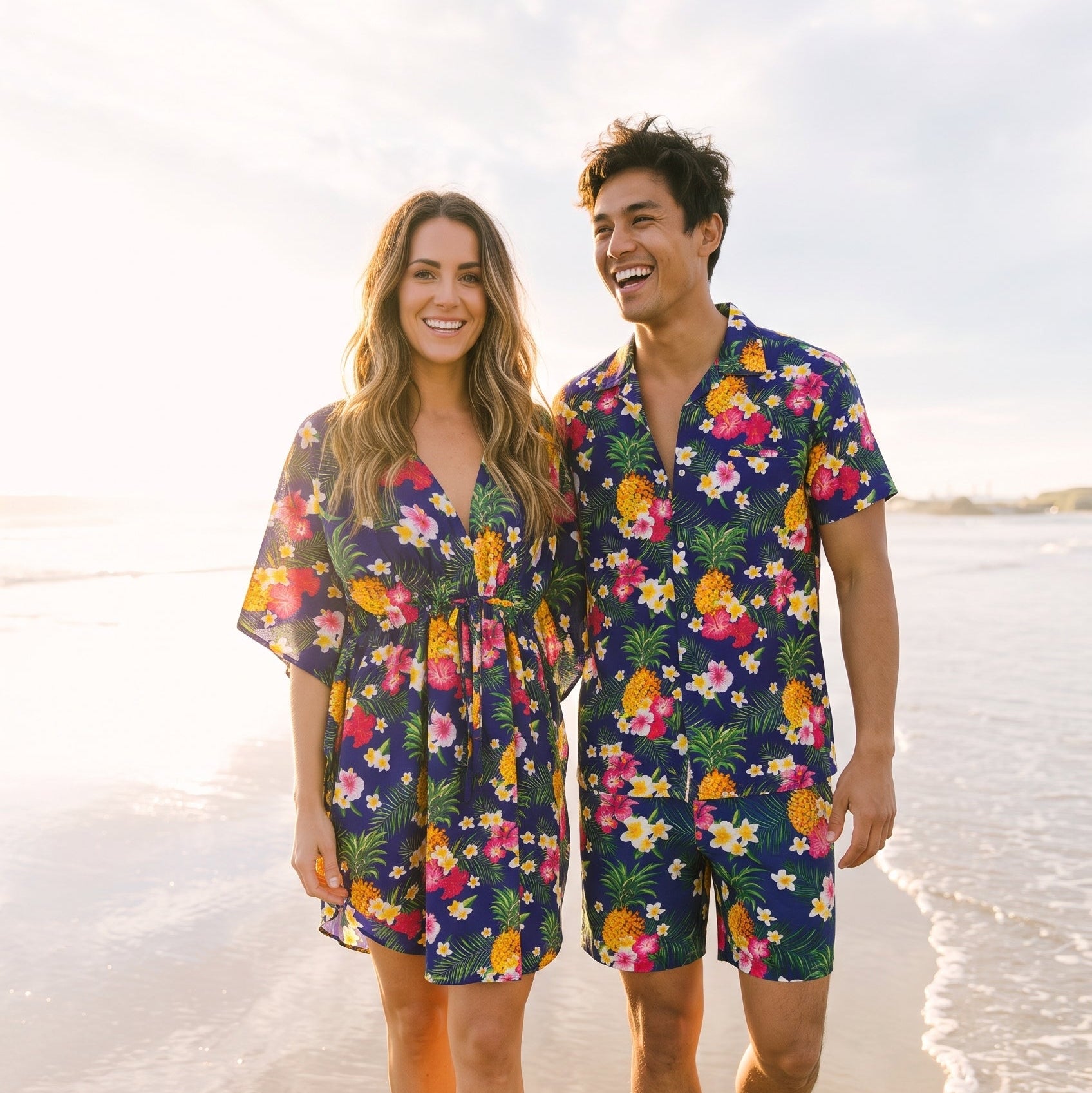 A smiling couple walking barefoot on a sandy beach wearing matching tropical floral-print swim cover-ups and shorts sets. The navy blue sets feature a vibrant multicolor print of pineapples, pineapples, and tropical flowers, with the woman's set showing a yellow pineapple detail on the front.
