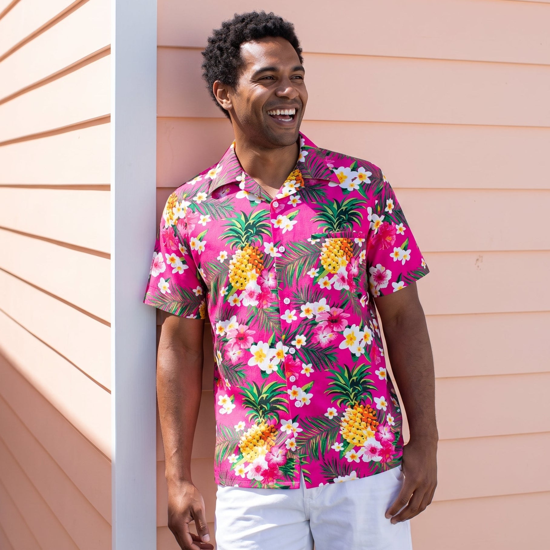 A man wearing a bright pink short-sleeve button-up shirt with a tropical pineapple and white flower print, paired with white pants, standing against a peach-colored house exterior.