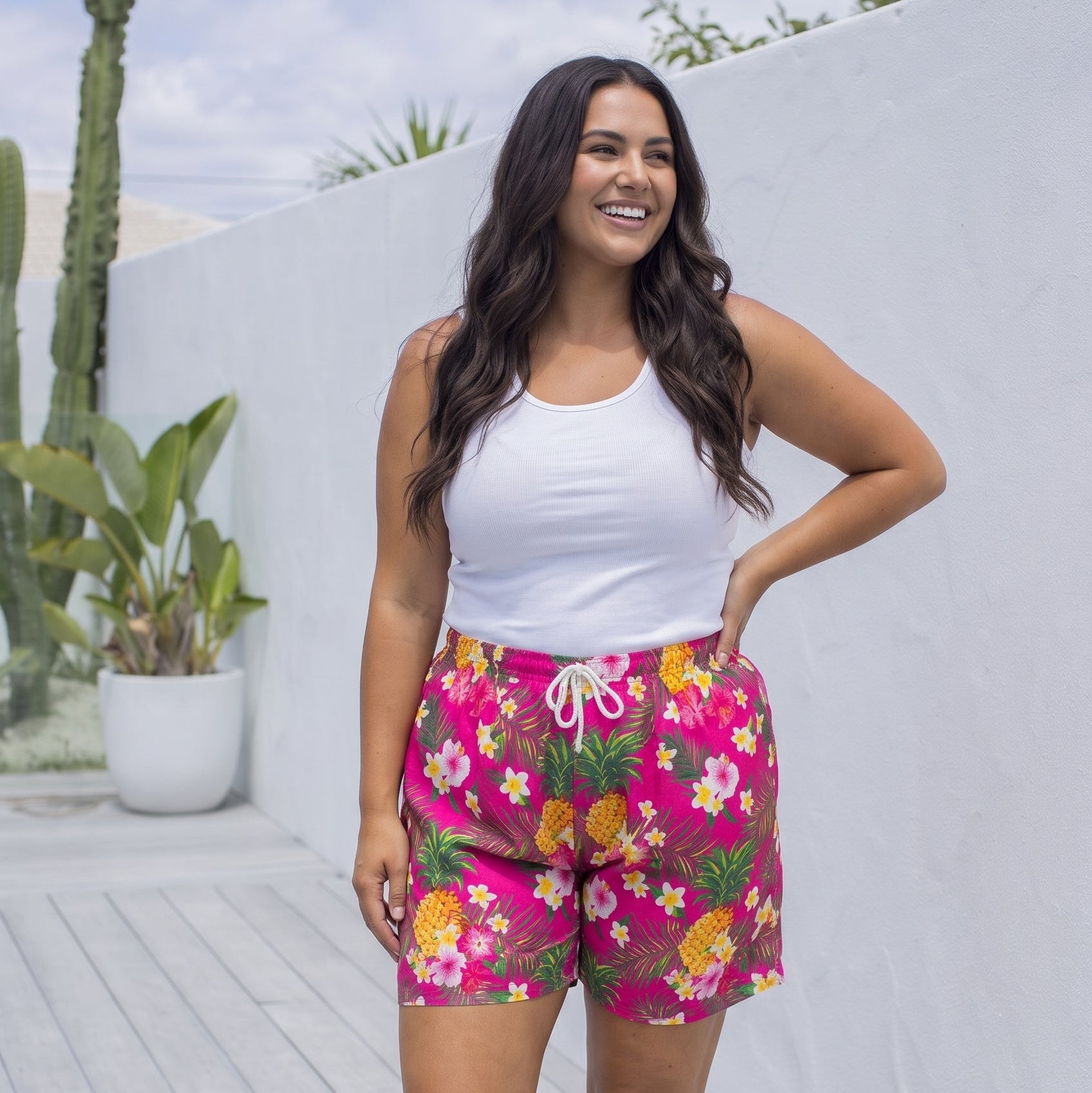 A woman modeling bright pink floral shorts with a white tank top, standing on a light wooden deck against a white wall.