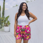 A woman modeling bright pink floral shorts with a white tank top, standing on a light wooden deck against a white wall.