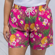 A woman modeling bright pink floral shorts with a white tank top, standing on a light wooden deck against a white wall.