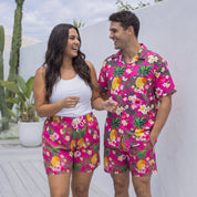 A man and a woman standing on a light gray wooden deck wearing matching tropical print swim shorts and a matching short-sleeve button-up shirt with a pineapple pattern, against a white wall background.