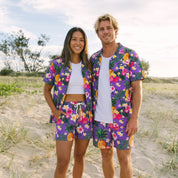 A man and a woman standing on a sandy beach wearing matching tropical floral print lounge sets with purple bases and multicolor pineapple, hibiscus, and orange patterns.