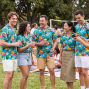 Five young adults standing together on a grassy outdoor lawn, wearing matching turquoise short-sleeve button-up shirts with a colorful tropical floral print and light shorts or a skirt. One person on the right is holding a phone, and drinks are visible in their hands.