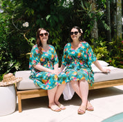Two women sitting side by side on a wooden lounge daybed outdoors by a pool, wearing matching teal and orange floral kimono-style dresses. Lush tropical greenery surrounds the patio setting.