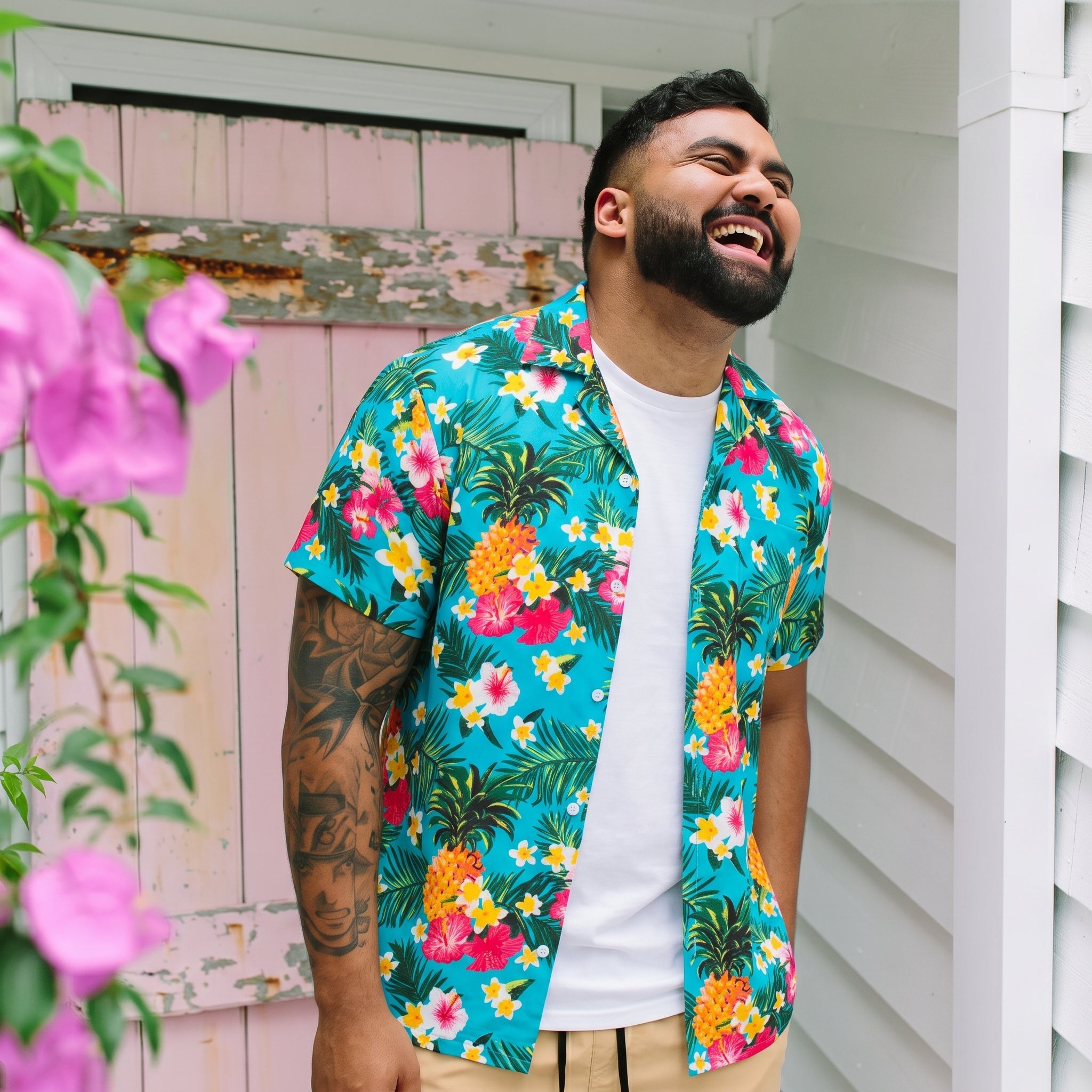 A man wearing a vibrant turquoise Hawaiian button-down shirt with a colorful tropical floral print, standing outdoors in front of a white house wall and a pink wooden door.