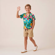 A young boy smiling and waving while modeling a vibrant tropical floral short-sleeve button-up shirt, tan shorts, and white sneakers against a solid light beige studio background.