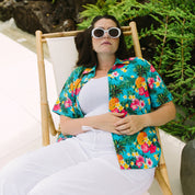 A woman reclining in a bamboo lounge chair outdoors, wearing a bright turquoise Hawaiian shirt with a colorful tropical floral print over a white tank top and white pants.