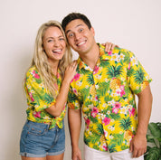 A smiling couple posing together in matching bright yellow tropical Hawaiian shirts with green palm leaves and pink floral prints, set against a plain light background.