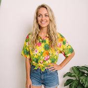 A woman with long blonde hair stands against a light wall wearing a bright yellow short-sleeve button-up shirt with a tropical floral and leaf print, tied at the waist, paired with denim shorts.