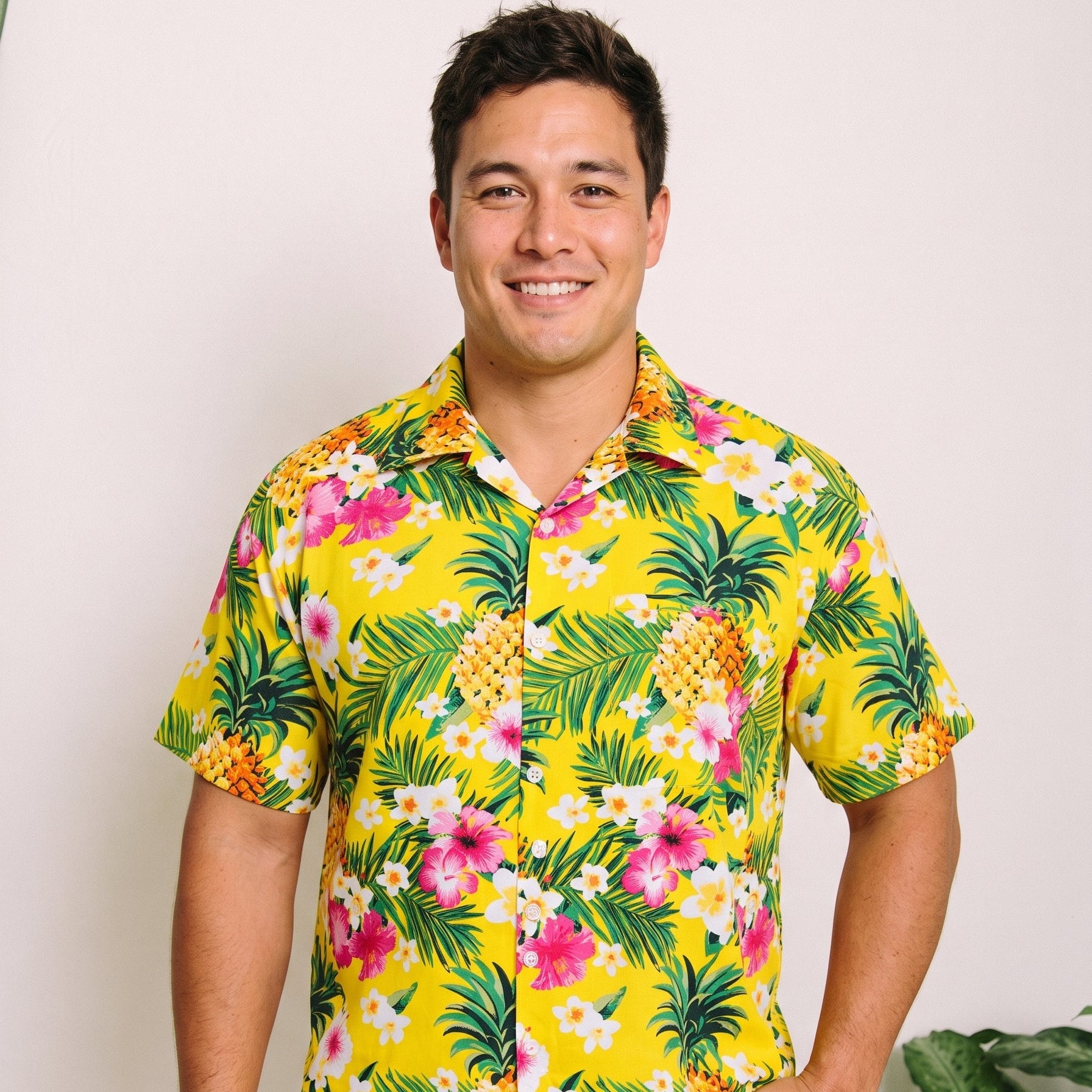 A man wearing a bright yellow short-sleeve button-up shirt with a tropical print of pineapples, palm leaves, and pink and white flowers, paired with white pants, standing against a light background.