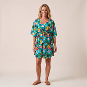 A woman modeling a short-sleeve V-neck dress with a colorful tropical floral and palm leaf print, tied at the waist, against a solid beige studio background.