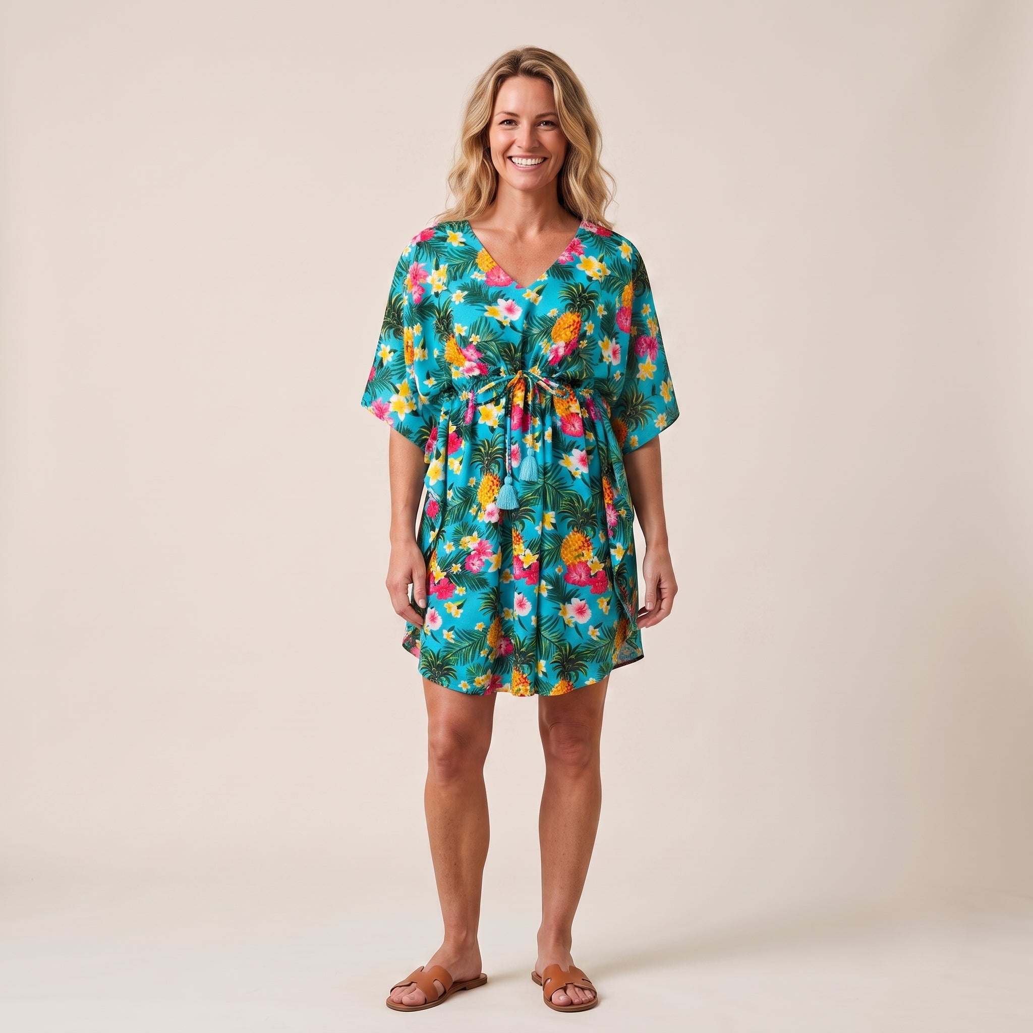 A woman modeling a short-sleeve V-neck dress with a colorful tropical floral and palm leaf print, tied at the waist, against a solid beige studio background.
