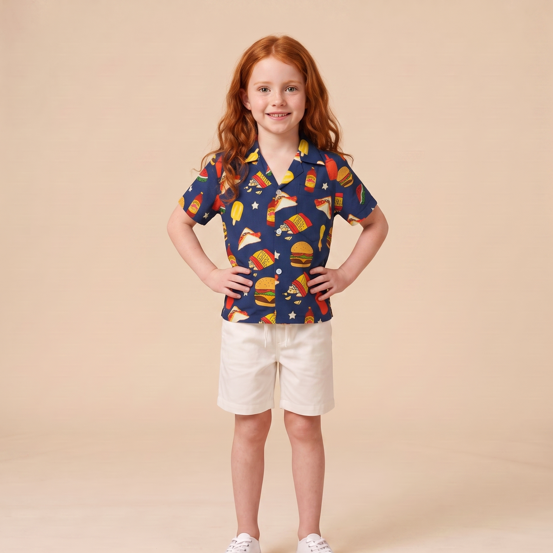 A young girl with red hair stands against a solid beige studio background, wearing a navy blue short-sleeve button-up shirt with a colorful fast-food print (burgers, hot dogs, pizza, fries) and cream shorts.