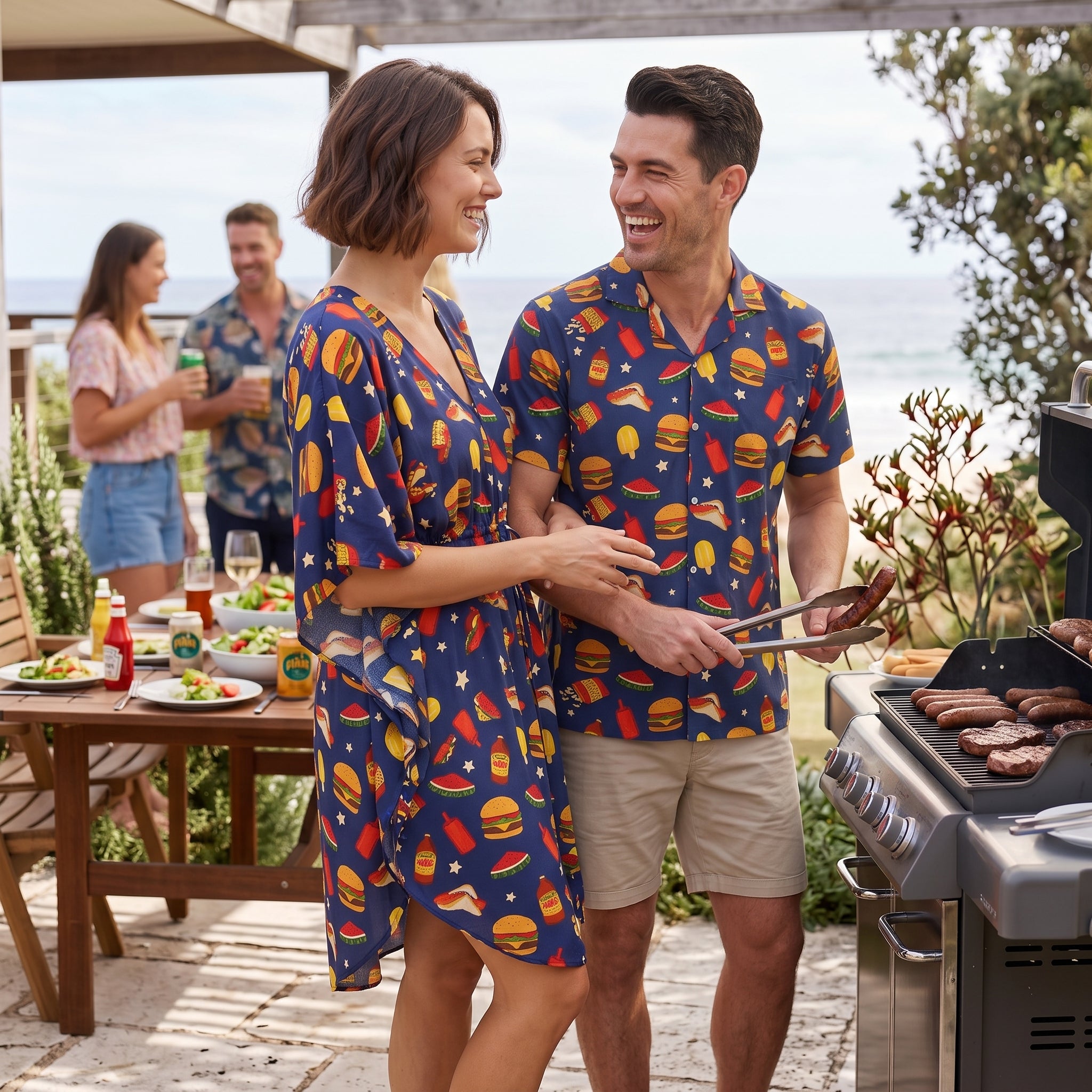A man and woman standing together at an outdoor patio grill, both wearing matching navy blue short-sleeve button-up shirts covered in a colorful hamburger print. The woman wears a knee-length matching dress, and the man wears khaki shorts.