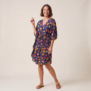 A woman modeling a navy blue kaftan dress with a colorful burger print, standing against a plain beige studio background.