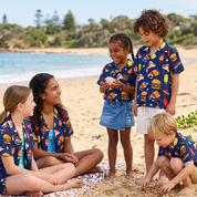 Five children wearing matching navy blue short-sleeve button-up shirts with a colorful burger and pizza print are playing on a sandy beach. The shirts feature a pattern of yellow cheese, red pepperoni, and green lettuce on a dark blue background.