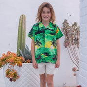 A young boy stands barefoot on a wooden deck wearing a vibrant green tropical print short-sleeve button-up shirt and off-white shorts, set against a white brick wall with potted cacti.