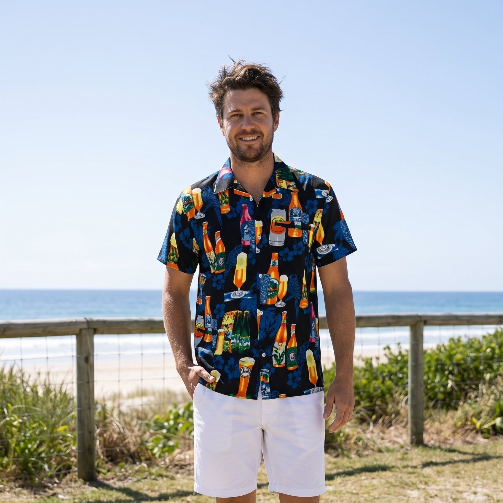A man wearing a navy blue short-sleeve button-down shirt with a colorful tropical beach-themed print, paired with white shorts and black sandals, standing on a beach path with the ocean and dunes in the background.