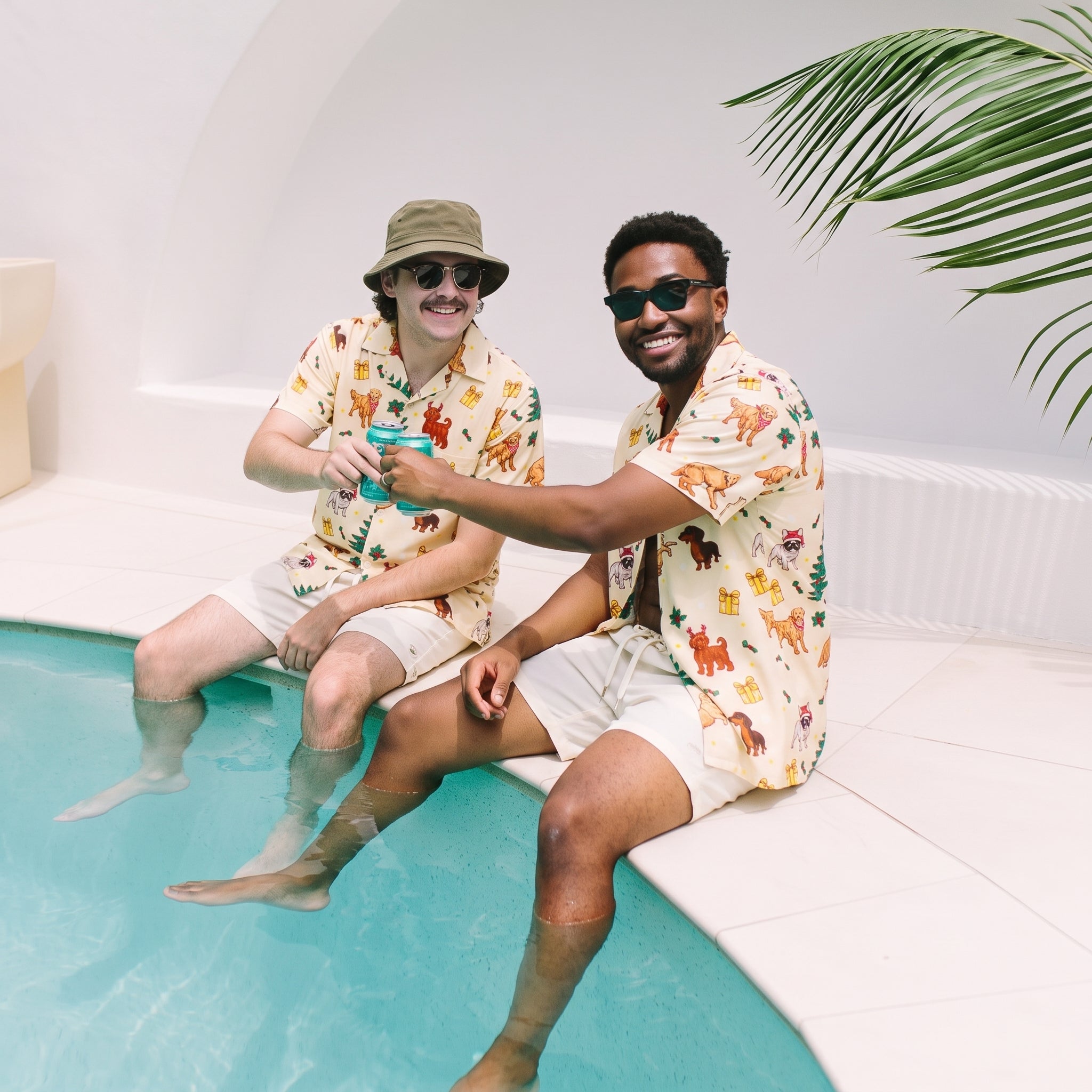 Two men sitting by a swimming pool wearing matching cream-colored short-sleeve button-up shirts with a colorful cartoon animal print and white shorts.