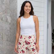 A woman modeling a white ribbed tank top and beige holiday-themed shorts with a Santa Claus and candy cane print, standing on a white porch.
