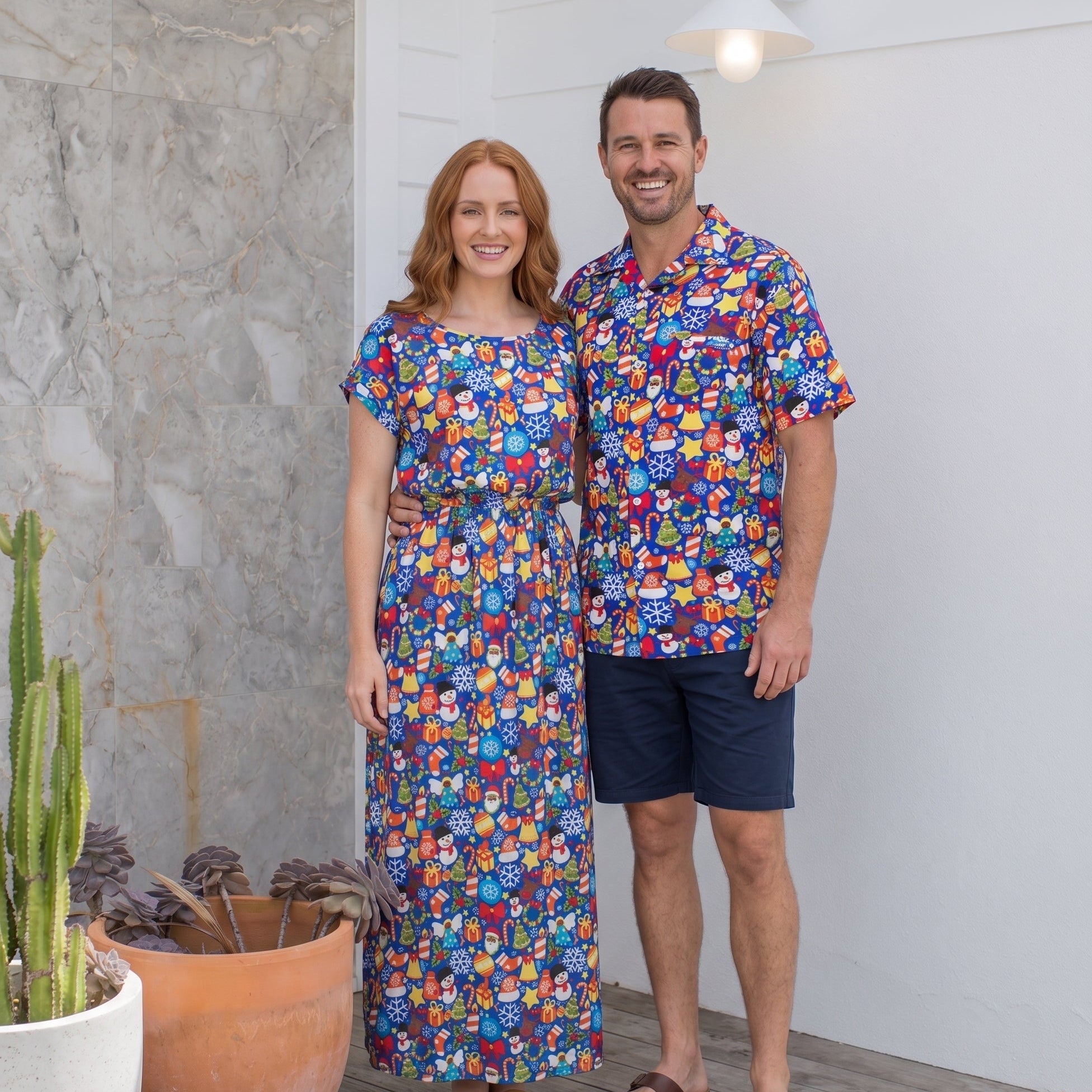 A man and a woman standing together on a porch wearing matching colorful short-sleeve button-up shirts and a matching maxi dress, with the man wearing navy shorts and sandals.
