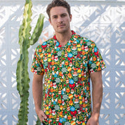 A man wearing a short-sleeve button-up shirt covered in a dense, colorful cartoon pattern of mushrooms, fruits, and characters, standing in front of a white geometric wall with a tall cactus to his left.