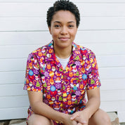A woman with short dark curly hair sits on stone steps in front of a white shiplap wall, wearing a short-sleeve button-up scrub top and matching shorts with a colorful cartoon pattern on a purple background.