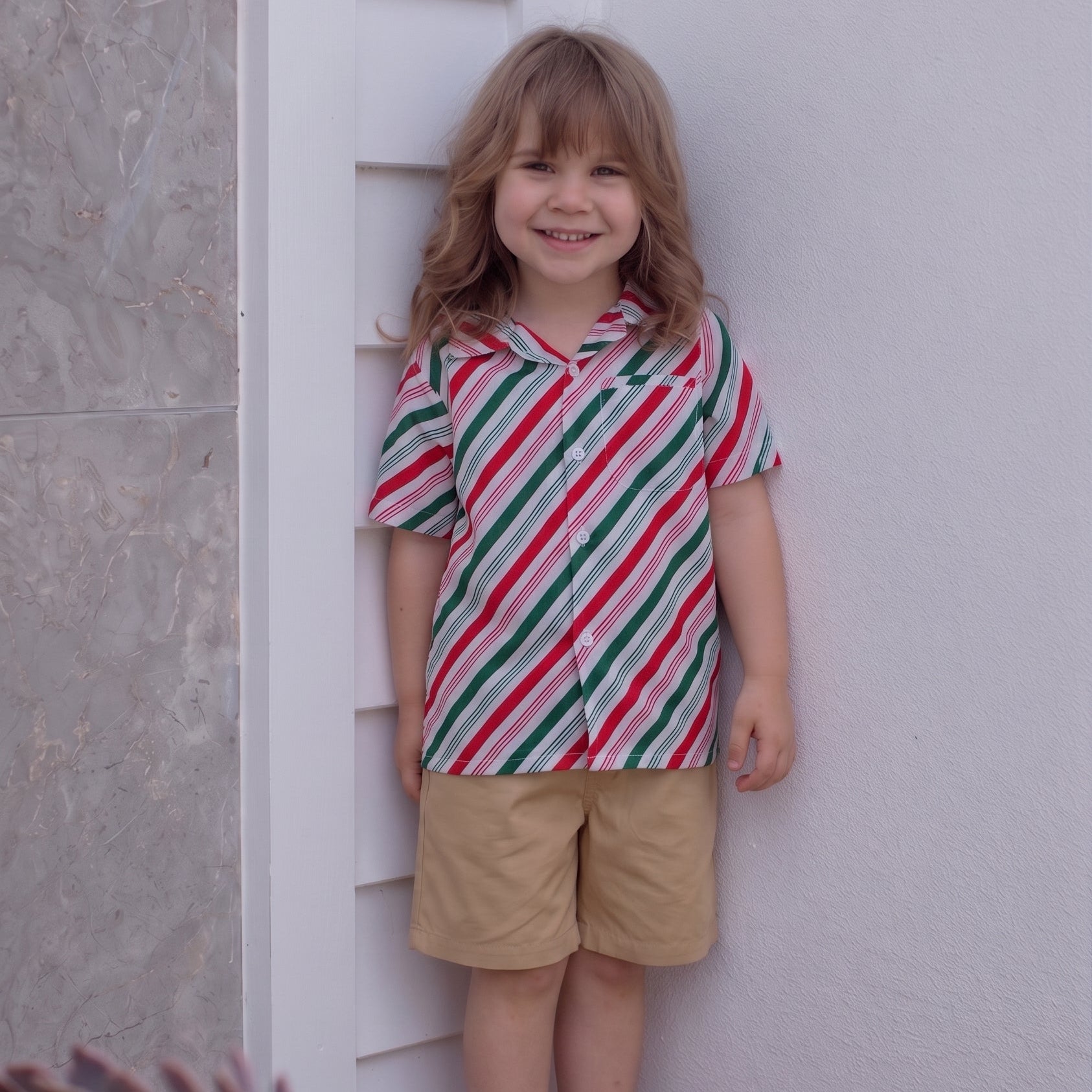 A young child standing barefoot against a white wall, wearing a short-sleeve button-up shirt with diagonal red, green, and white stripes and tan shorts.