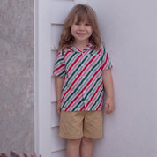 A young child standing barefoot against a white wall, wearing a short-sleeve button-up shirt with diagonal red, green, and white stripes and tan shorts.