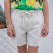A young boy stands barefoot on a wooden deck wearing a vibrant green tropical print short-sleeve button-up shirt and cream-colored shorts, set against a white brick wall with potted cacti.