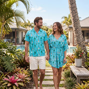 A man and a woman walking hand-in-hand on a tropical resort path, wearing matching turquoise holiday-themed short-sleeve shirts and a matching dress with a snowman and pine tree pattern.
