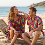 A man and a woman sitting on beach lounge chairs on a sandy beach, wearing matching colorful tropical floral short-sleeve shirts and a matching floral dress.