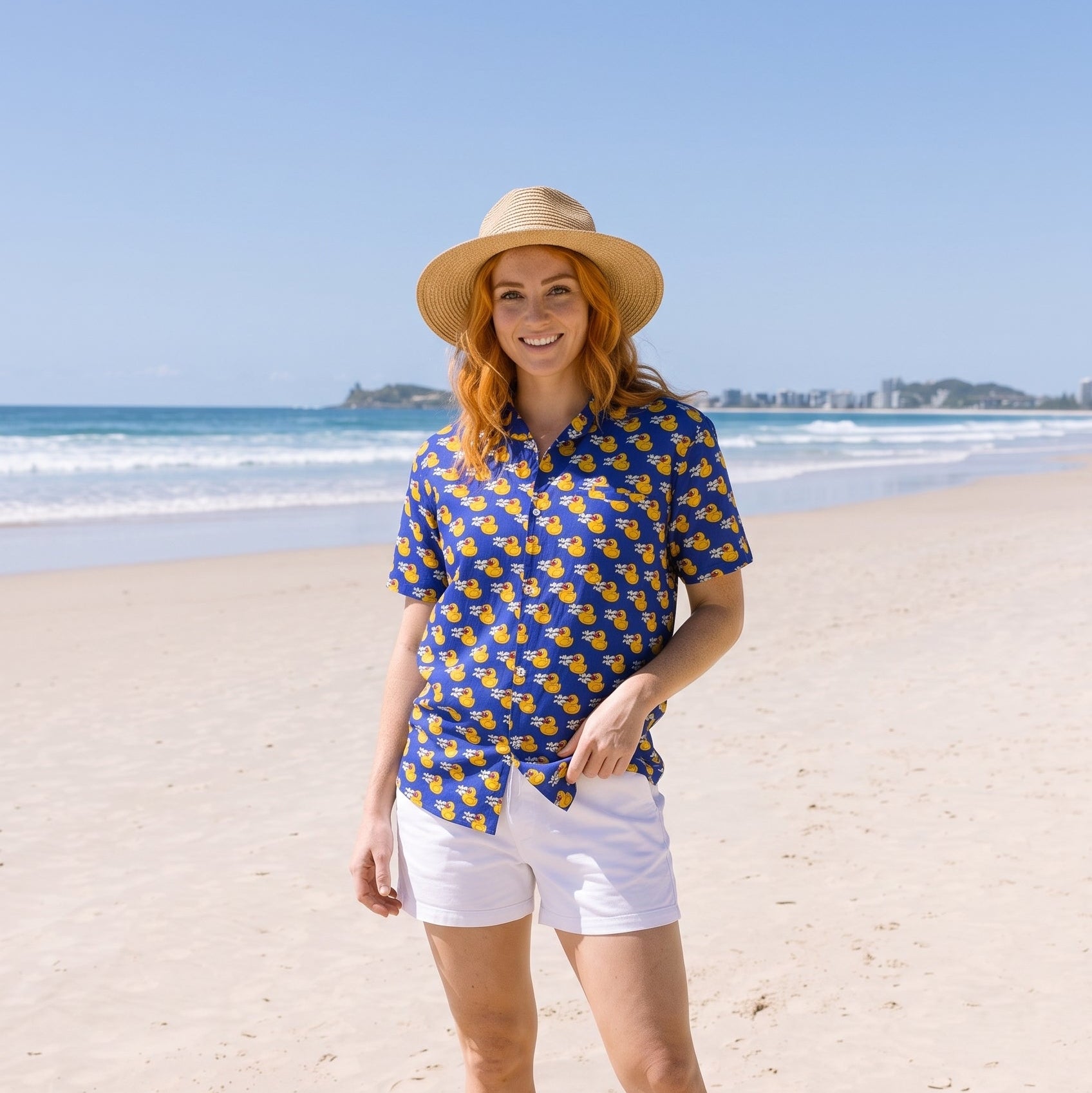 A woman stands on a sandy beach wearing a blue short-sleeve button-up shirt with a yellow pattern, white shorts, a straw hat, and black flip-flops, with the ocean and blue sky in the background.