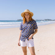 A woman stands on a sandy beach wearing a blue short-sleeve button-up shirt with a yellow pattern, white shorts, a straw hat, and black flip-flops, with the ocean and blue sky in the background.