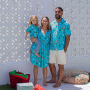 A family of three posing outdoors in matching holiday-themed matching sets. The mother wears a turquoise blue belted wrap dress with a Christmas tree and snowman pattern, holding a young girl in a matching blue dress.