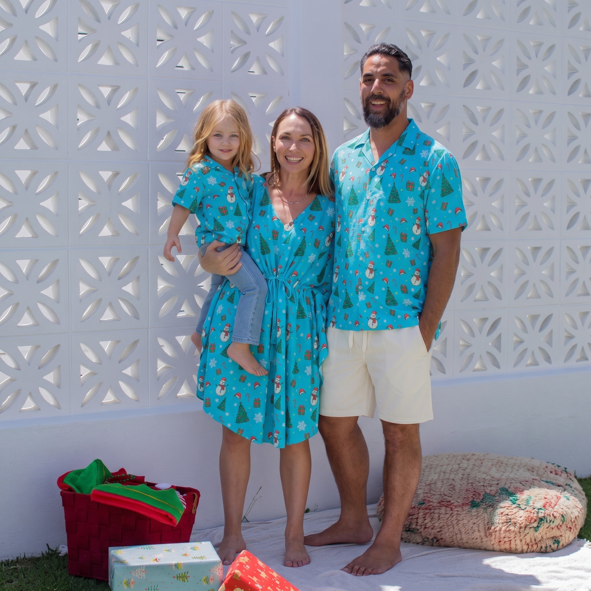 A family of three posing outdoors in matching holiday-themed matching sets. The mother wears a turquoise blue belted wrap dress with a Christmas tree and snowman pattern, holding a young girl in a matching blue dress.