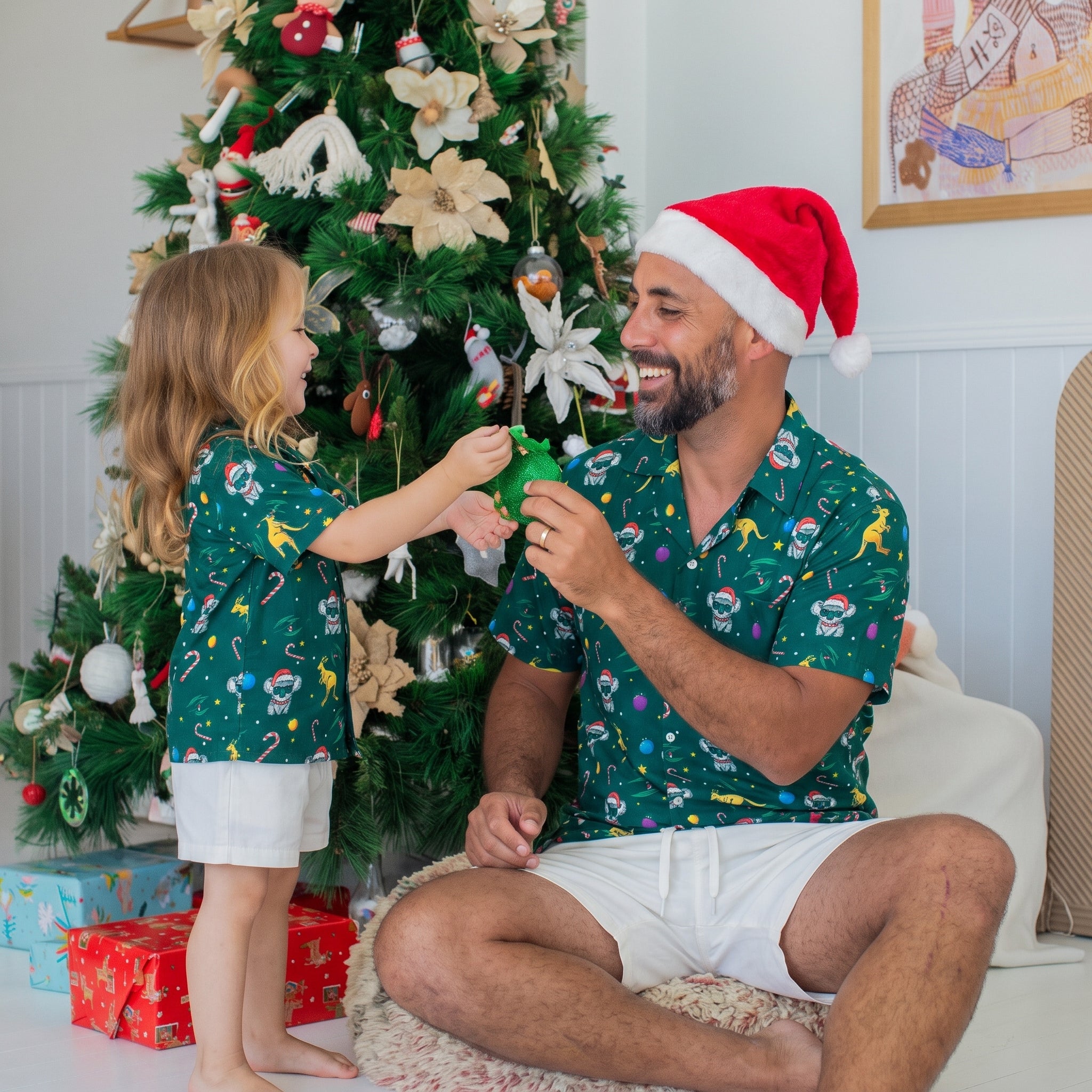 A man and a young girl wearing matching green Christmas-themed short-sleeve shirts sit together in front of a decorated Christmas tree, with the man wearing a red Santa hat and white shorts.