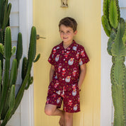 A young boy stands barefoot in front of a yellow door, wearing a matching short-sleeve pajama set with a red base and a festive Santa Claus snowman print. The scene is framed by tall potted cacti on both sides.